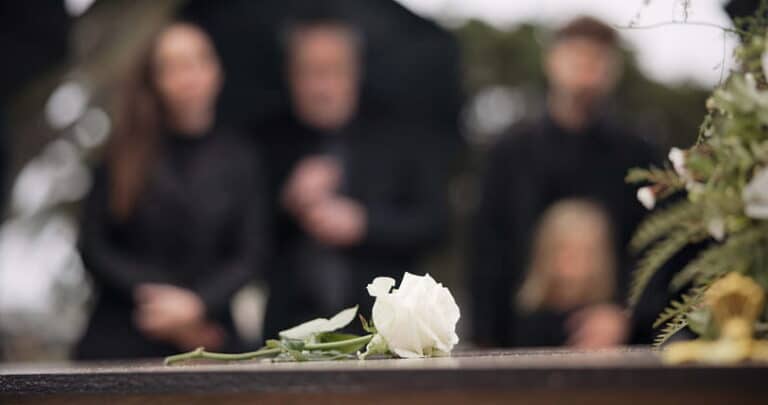 A white rose sitting on a gravesite with mourners blurred out in the background.